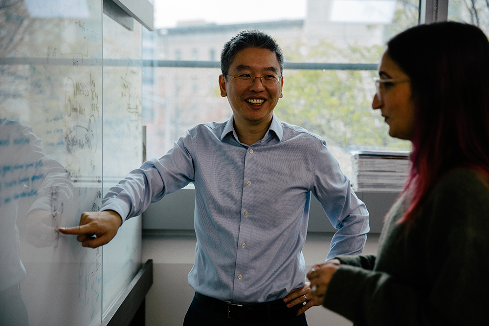 Harris Wang speaks with a lab member in his office, pointing to an equation on a whiteboard.